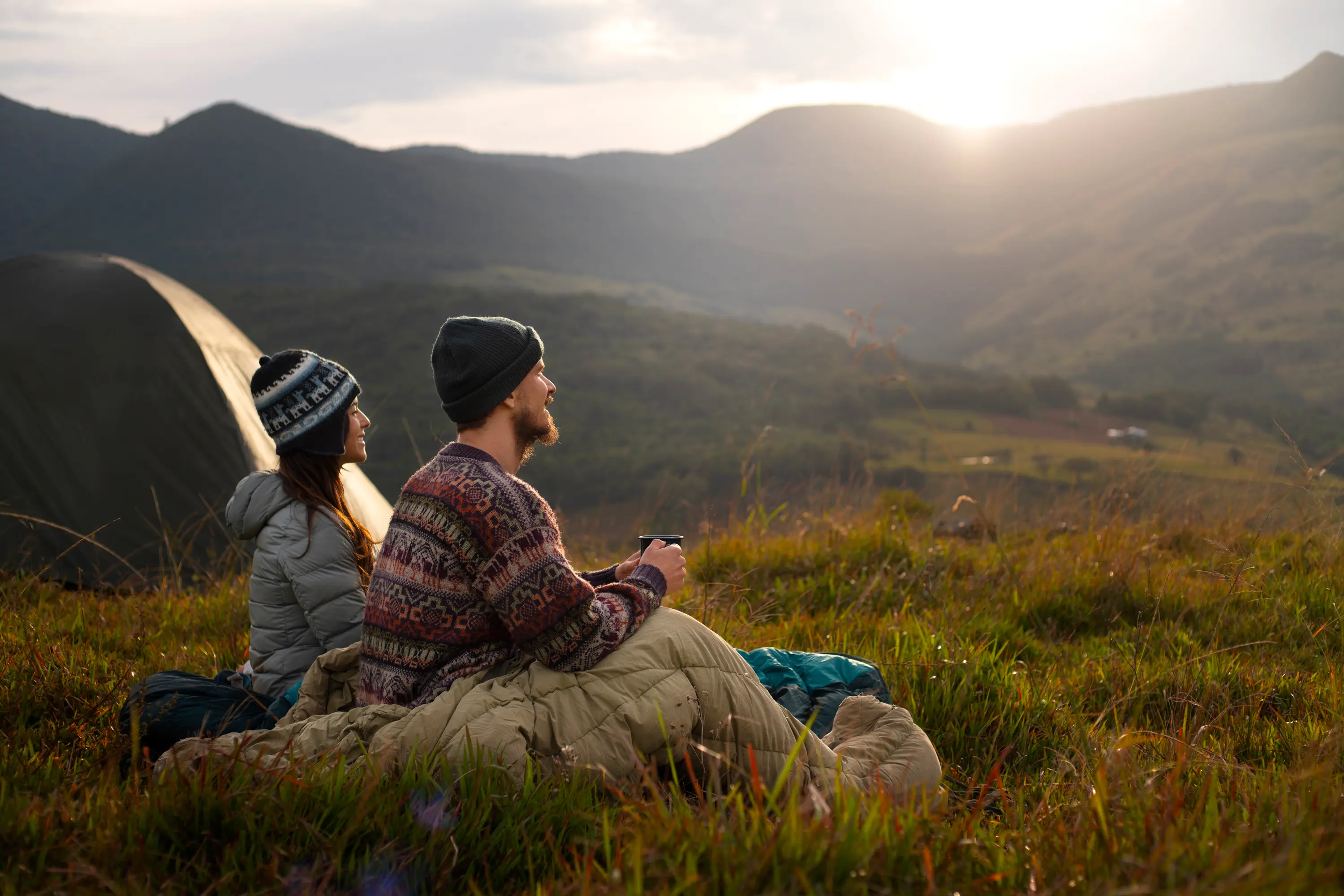 Two people sitting on grass near a tent at sunrise, enjoying the mountain view and drinking from a cup.