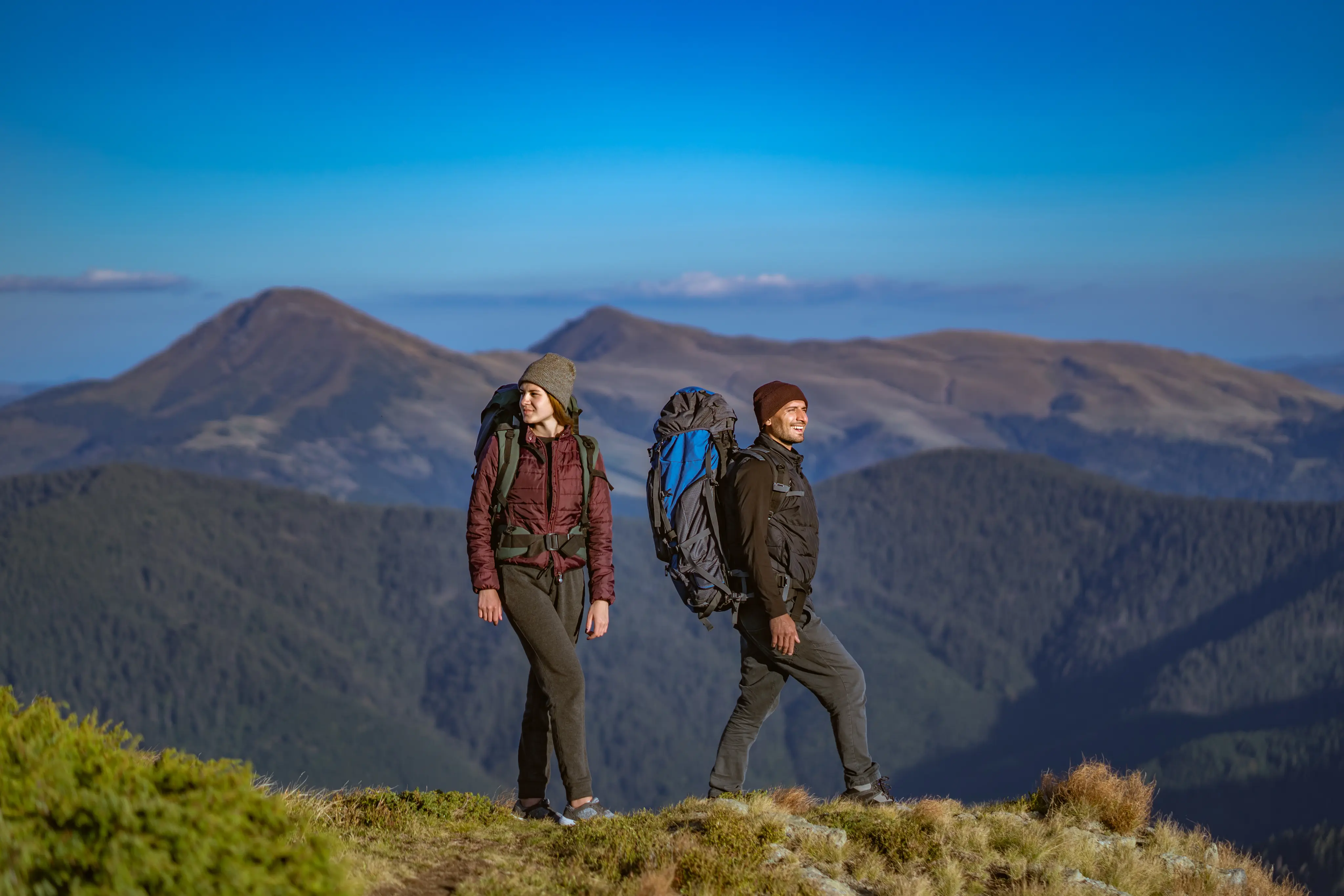 Two hikers with backpacks standing on a mountain ridge with rolling hills and clear blue sky in the background.
