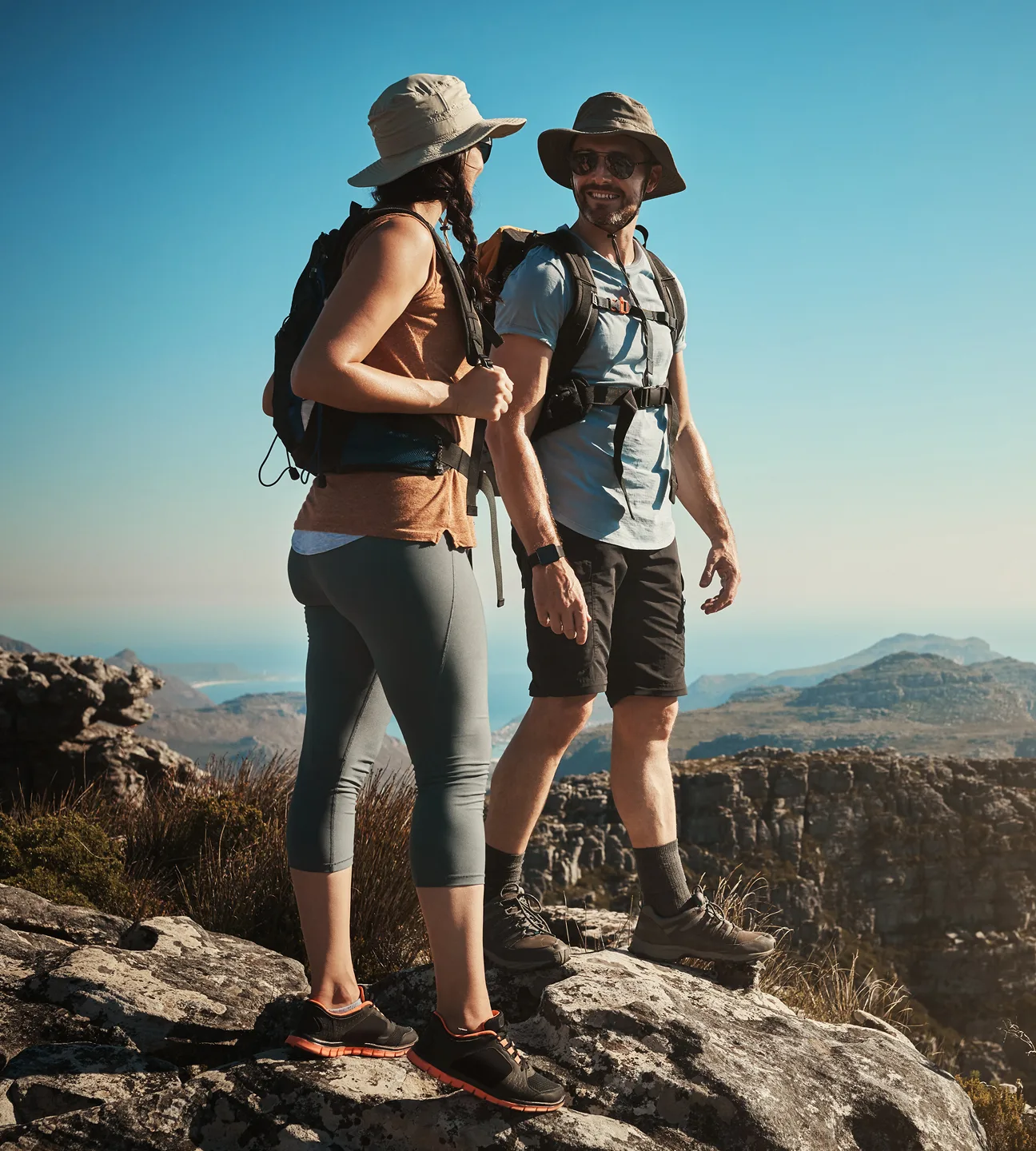 Two hikers with backpacks and hats standing on rocky terrain, overlooking a mountainous landscape under a clear blue sky.
