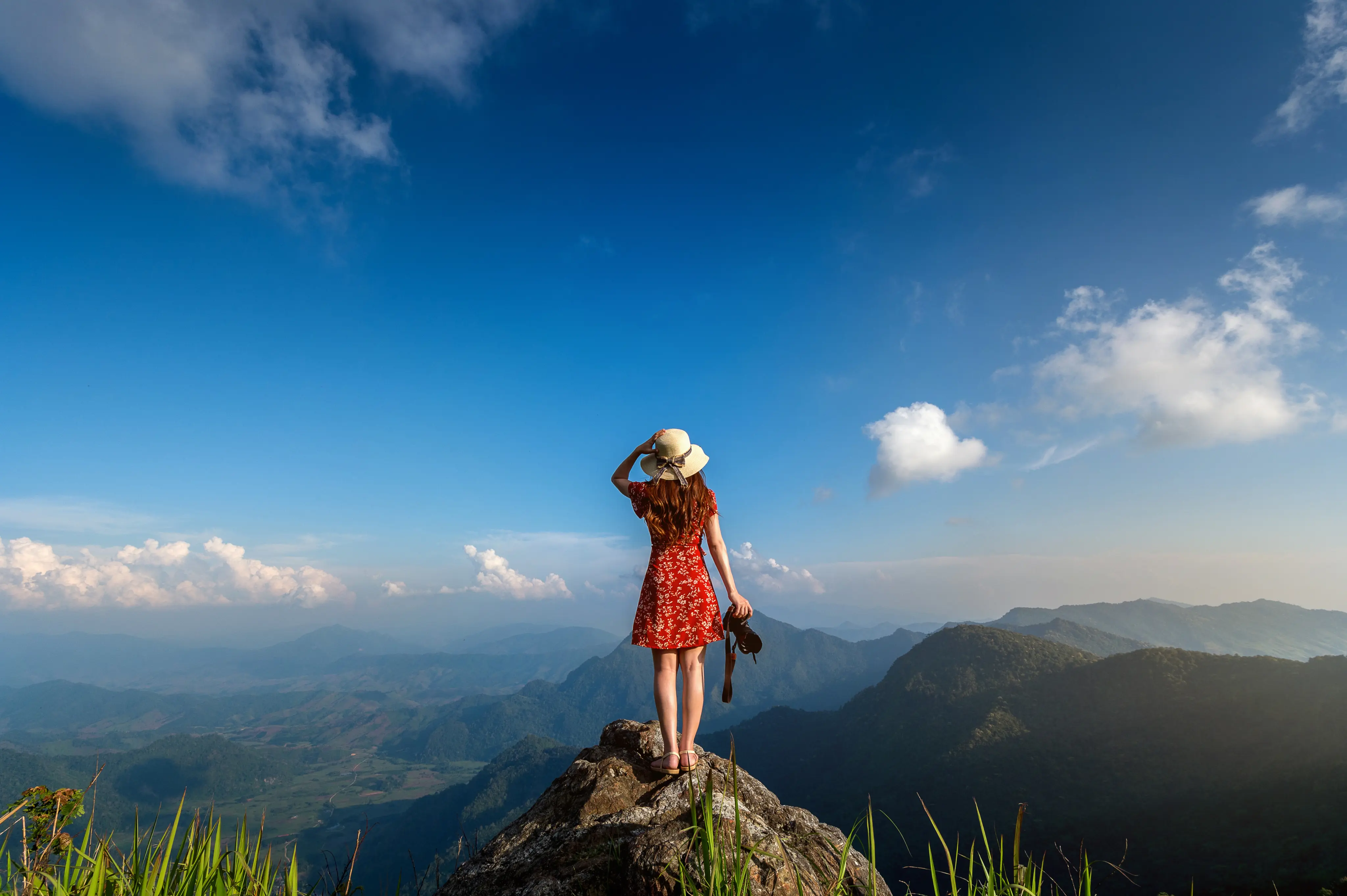Woman in a red dress and hat standing on a mountain peak holding a camera, overlooking a scenic valley and mountain range under a blue sky.
