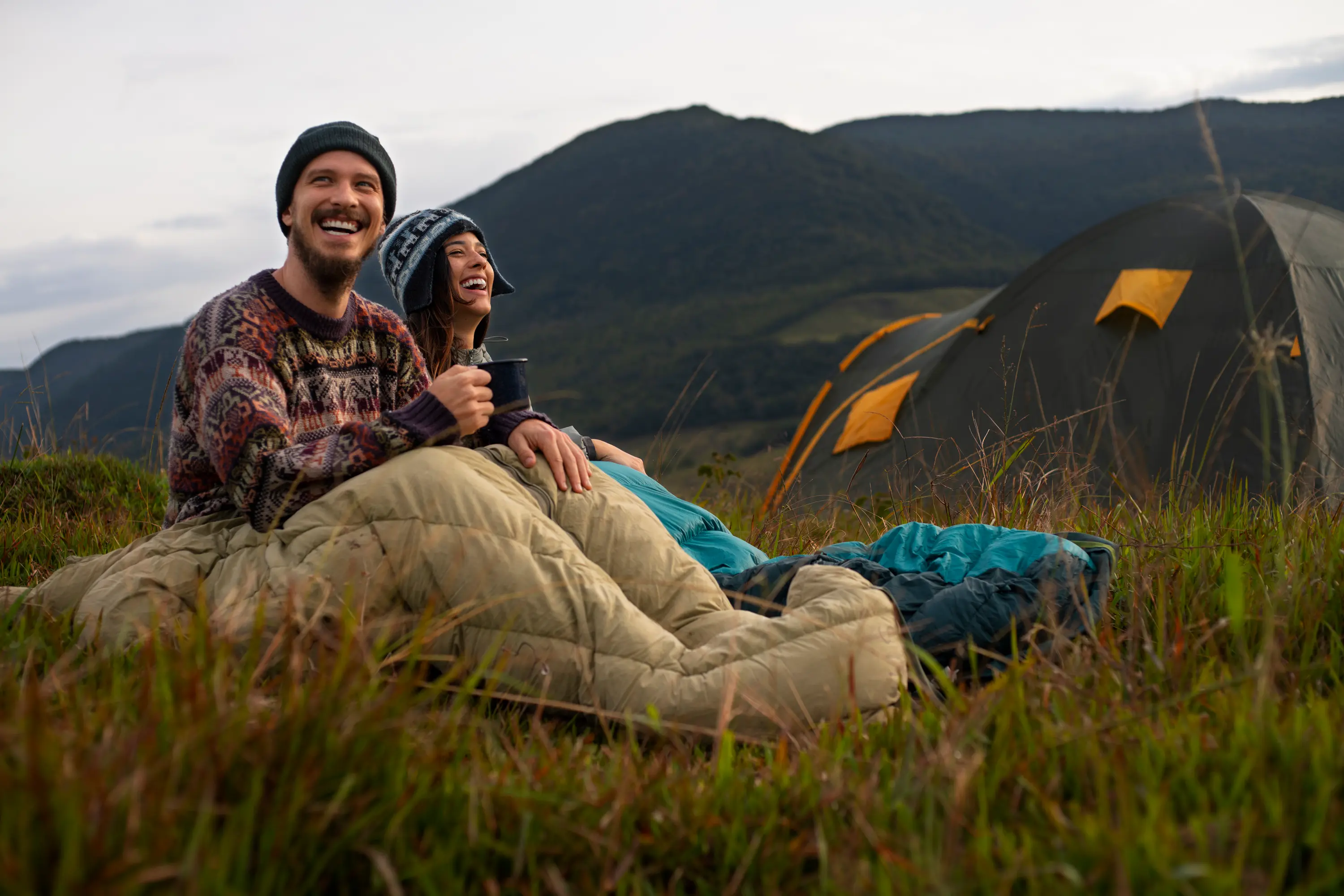 Smiling couple sitting in sleeping bags on grass near a tent with mountains in the background.