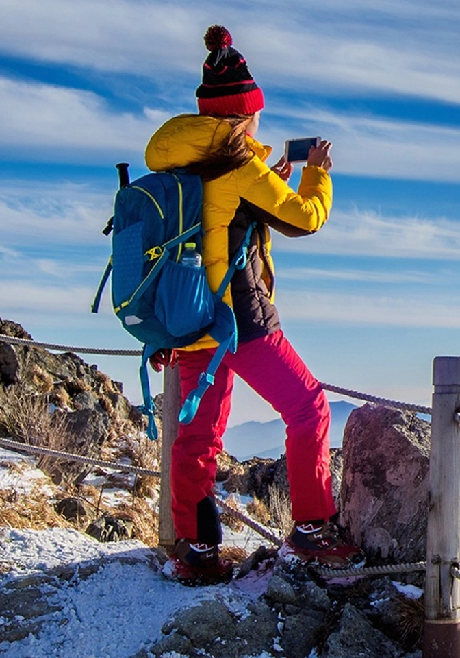 Hiker in yellow jacket and red pants with blue backpack taking a photo on a snowy rocky mountain trail.