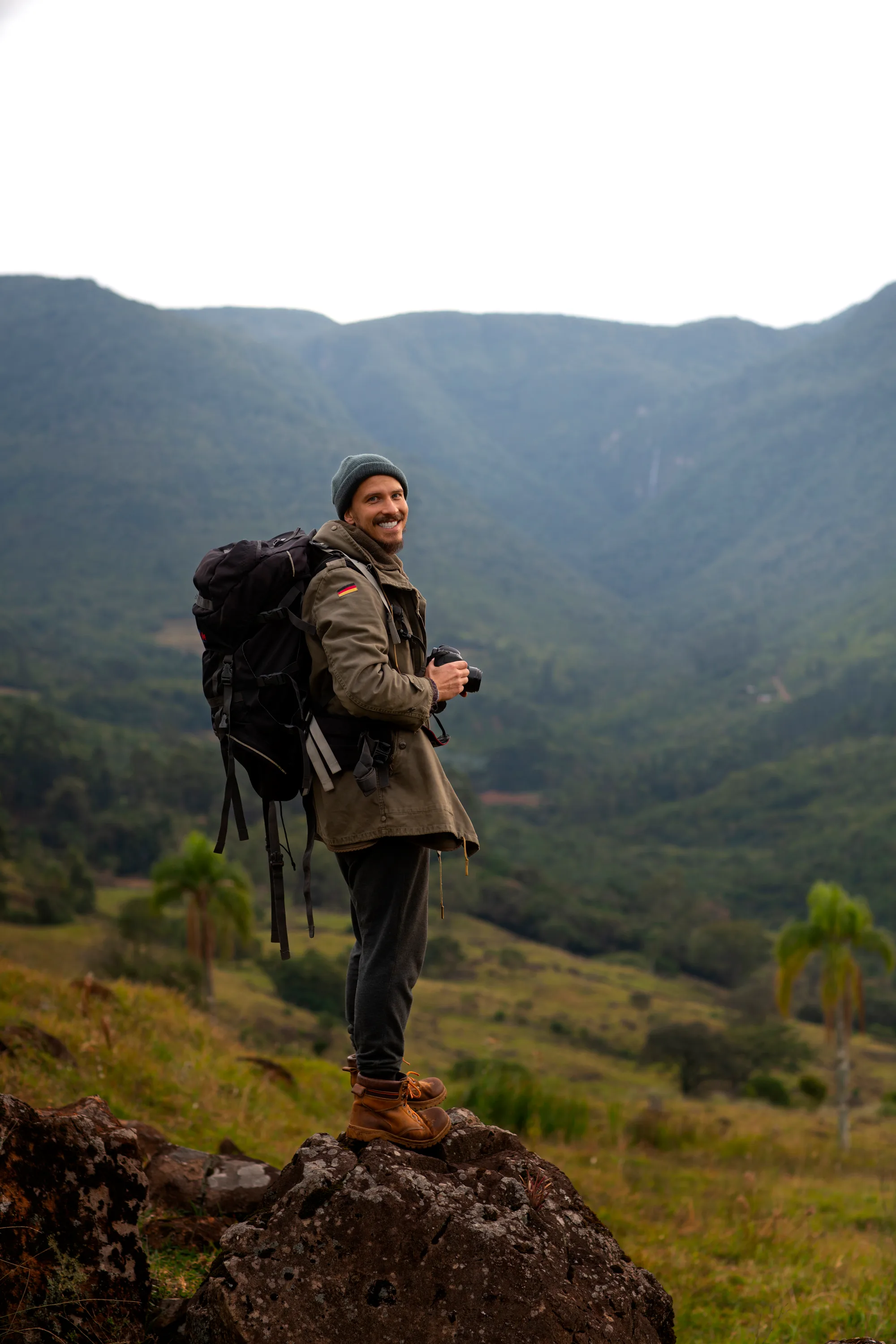 Smiling man wearing a beanie and backpack, standing on a rock with a camera in hand, overlooking a green valley with mountains.