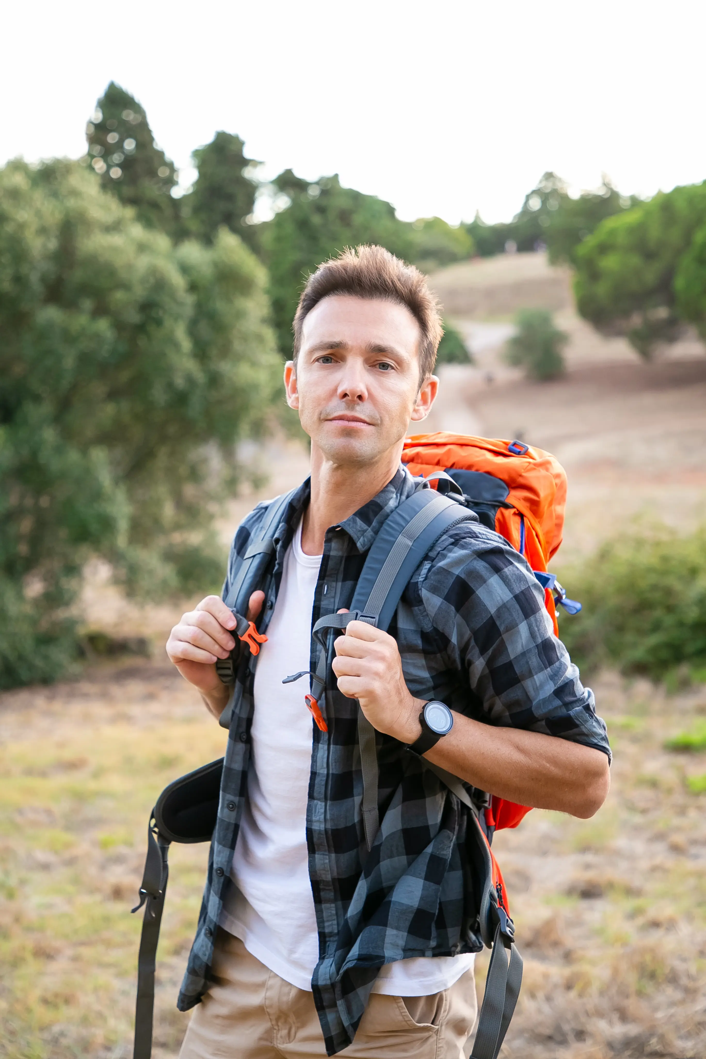 Man wearing a black checkered shirt and carrying an orange backpack outdoors in a natural setting.